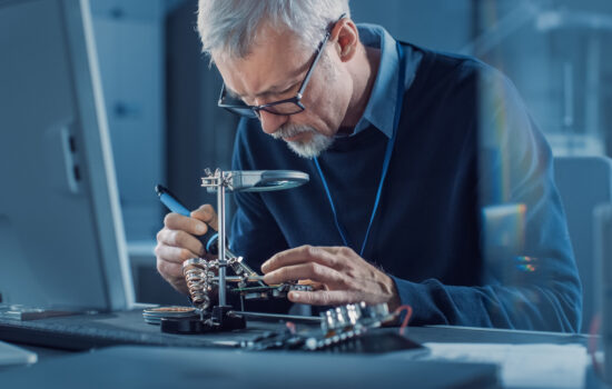 Electronics Maintenance Engineer Soldering Motherboard, Microchip and Circuit Board, Looking through Magnifying Glass, Consults Personal Computer. Electronics Repair and Testing